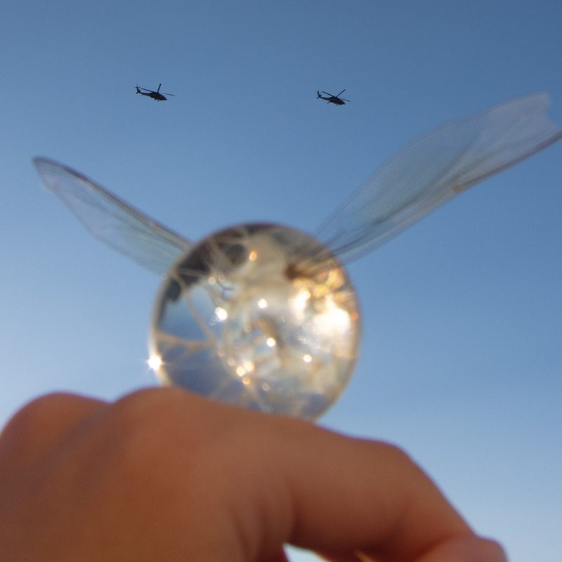 Glass orb with insect wings held up against a blue sky with two helicopters